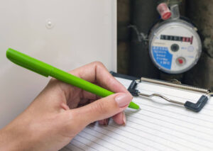 A close-up of a hand writing something on a clipboard with a water meter in the background.